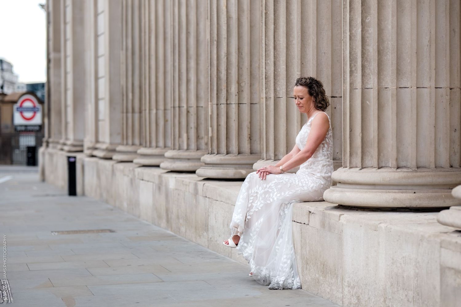 A bride in an elegant white lace dress poses between classical stone columns on a grand building exterior.