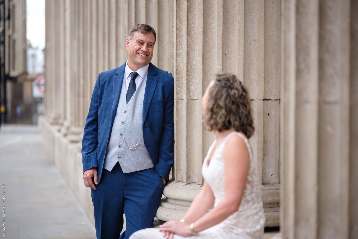Two people having a wedding photo session in formal attire near classic stone columns outdoors.