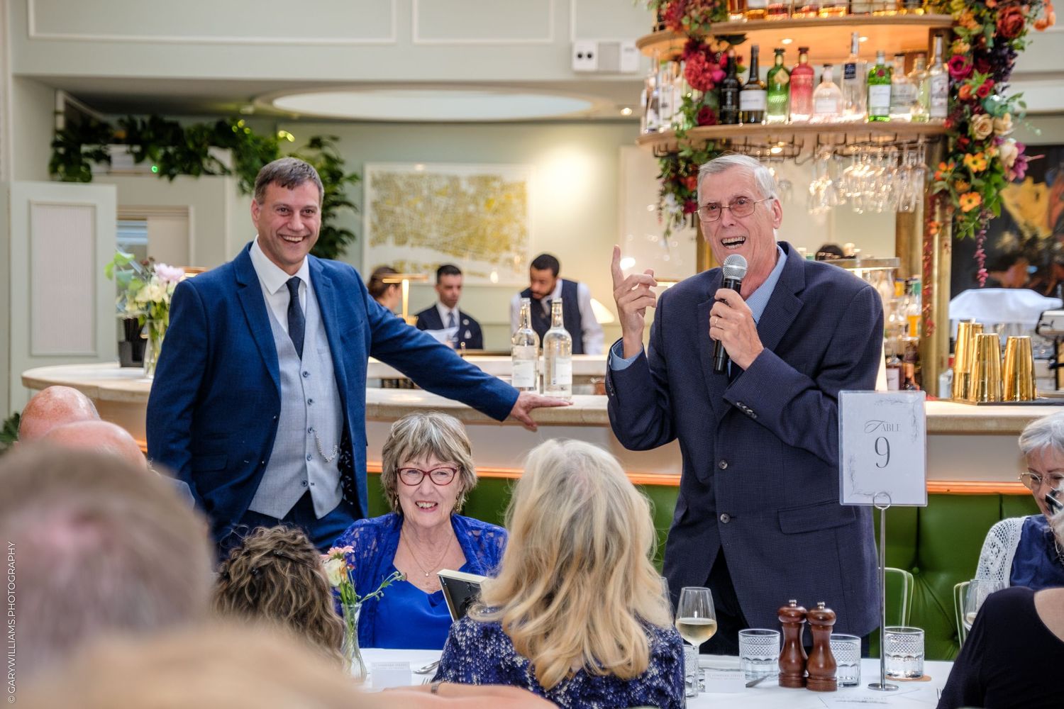 A group of people gather at an elegant event with floral decorations and formal attire in a bright indoor setting.