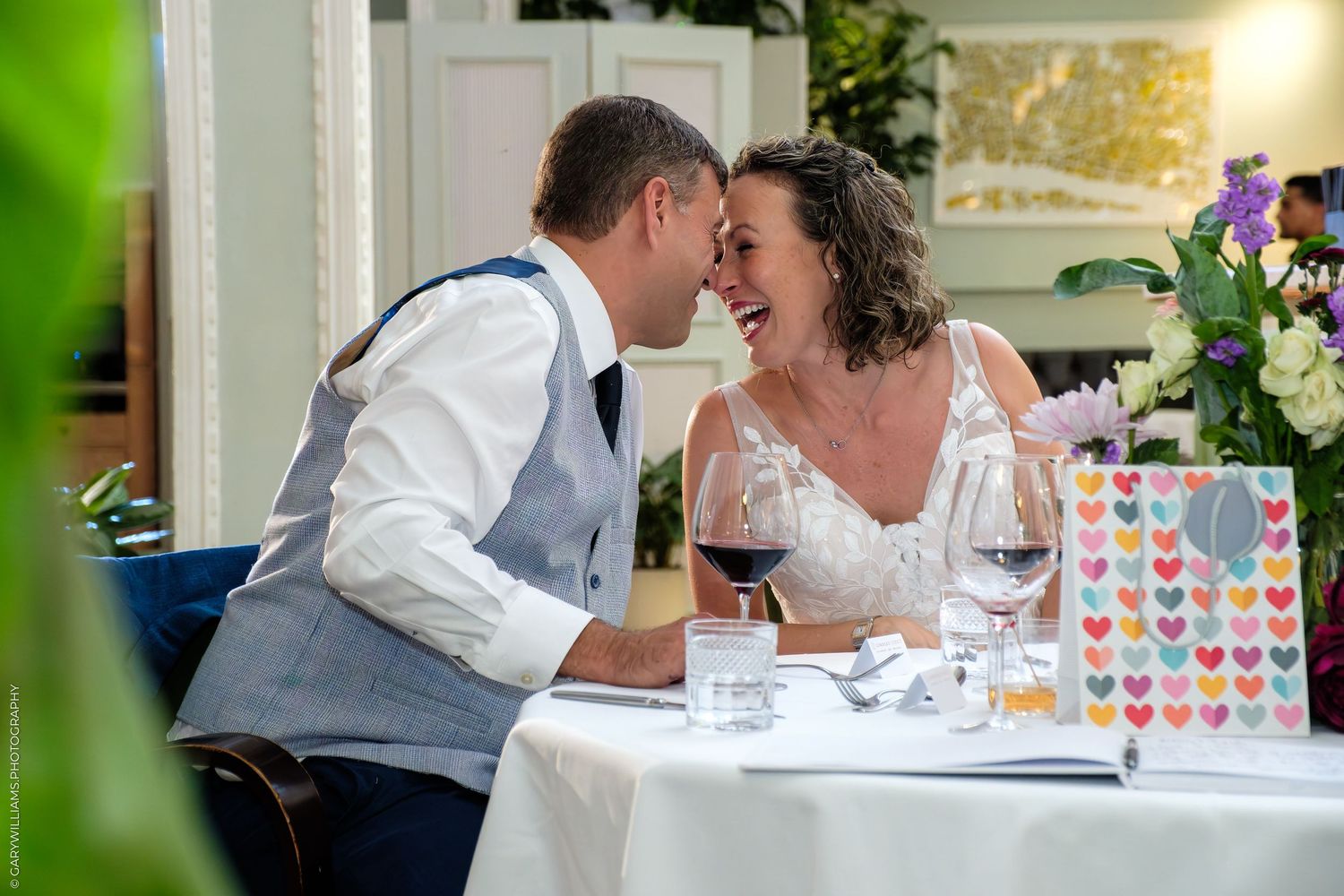 Two people share an intimate moment at a restaurant table with wine glasses and colorful heart decorations on display.