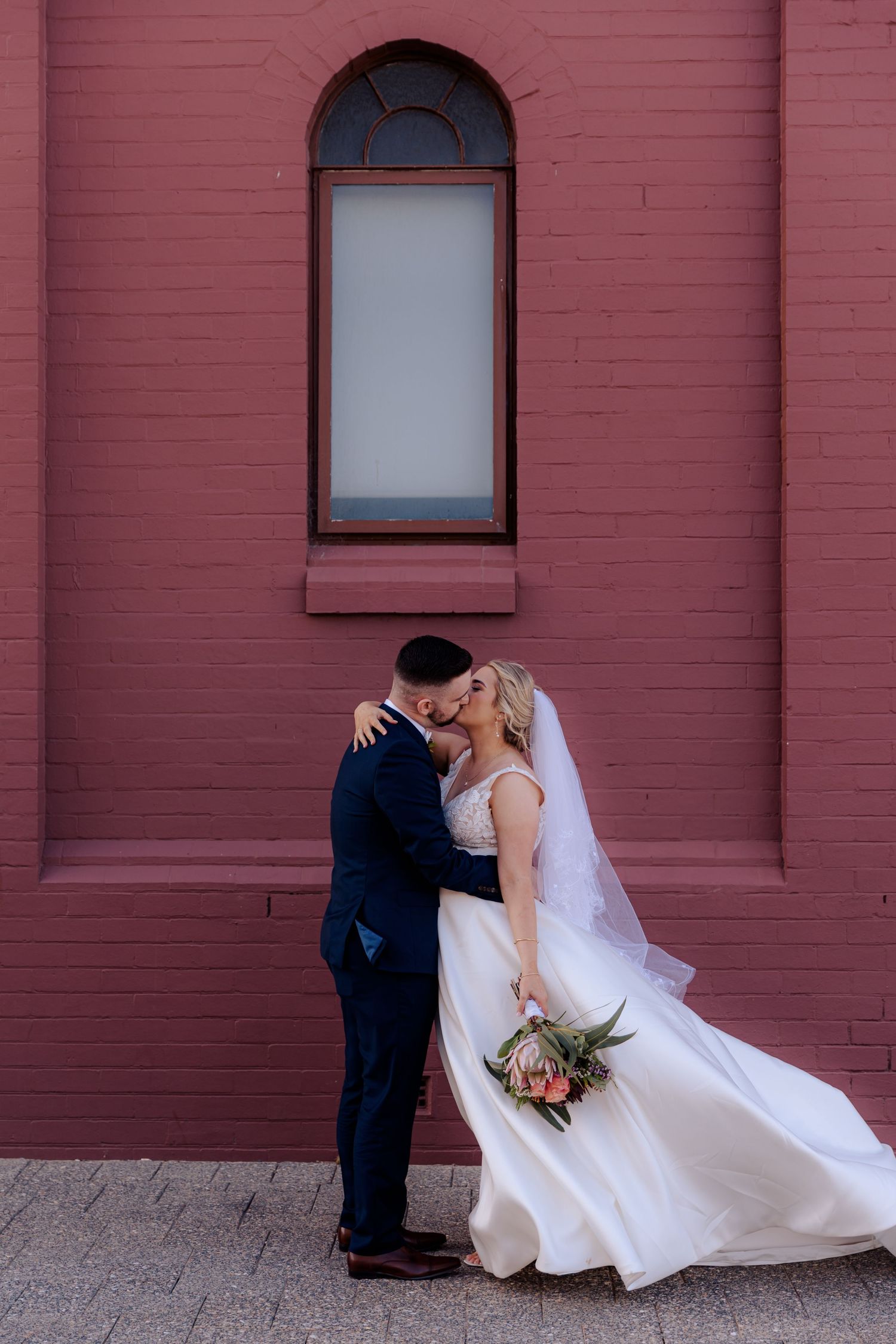 A couple shares romantic kiss in front of pink brick wall with wedding attire silhouetted against the backdrop.