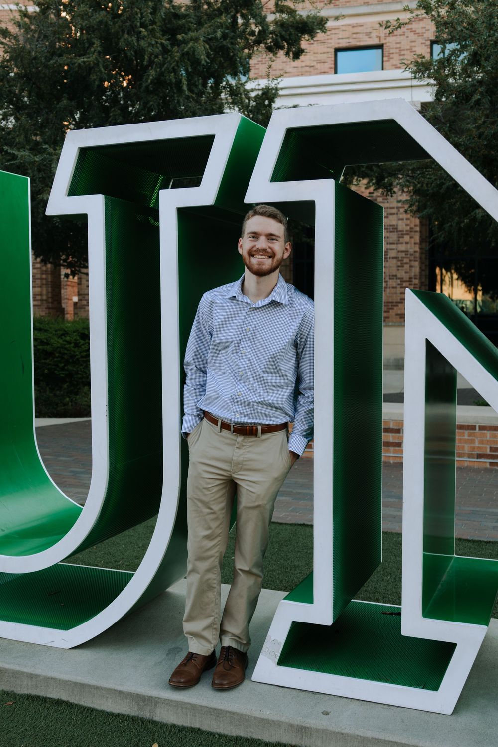 Graduate poses with large white and green university letters on campus lawn.