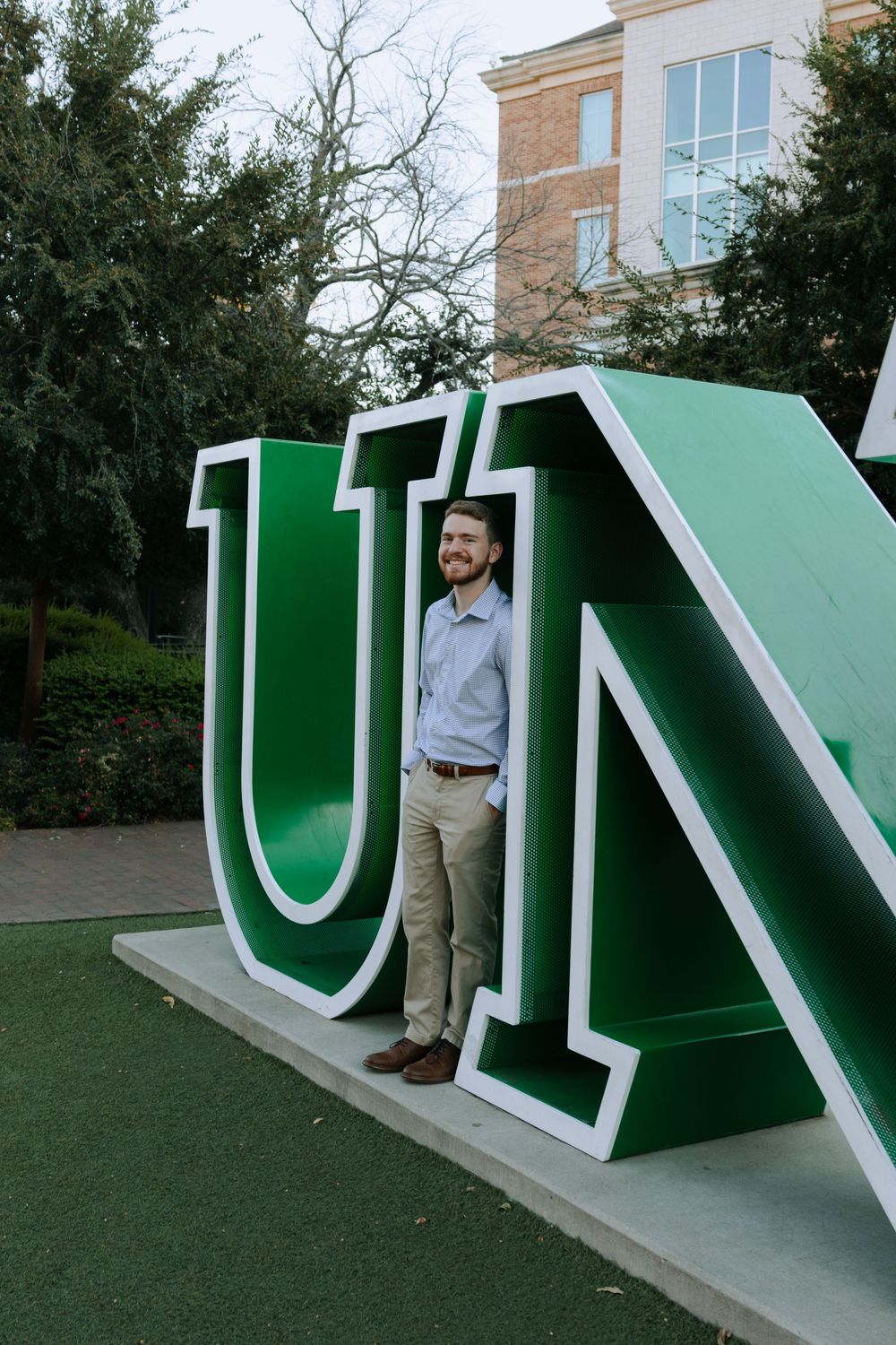 Student stands beside oversized white and green university sign on grassy area.
