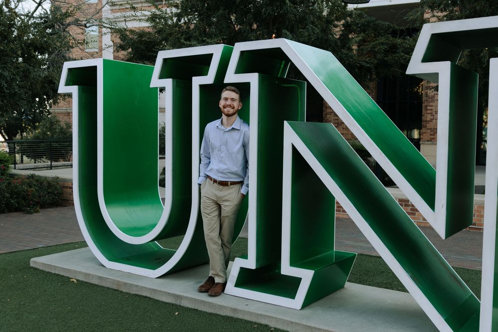 Graduate in green academic regalia poses next to large outdoor university sign.
