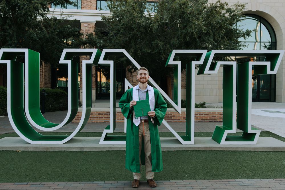 Graduate in casual attire poses near large green and white university signage outdoors.