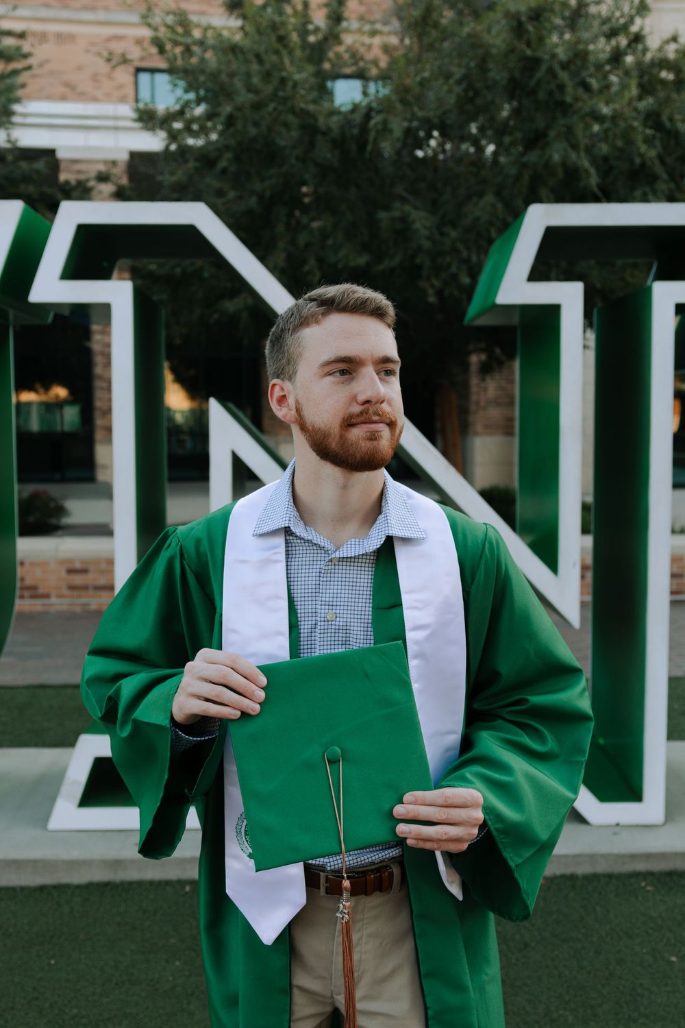 Graduate wearing green cap and gown holds diploma while standing outdoors.