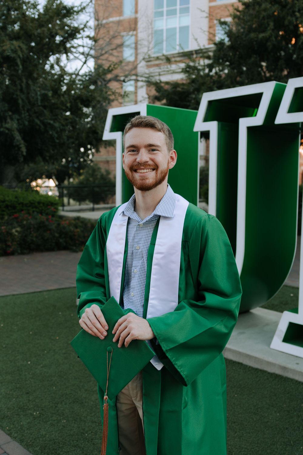 Graduate wearing green cap and gown with white stole poses by university letters.