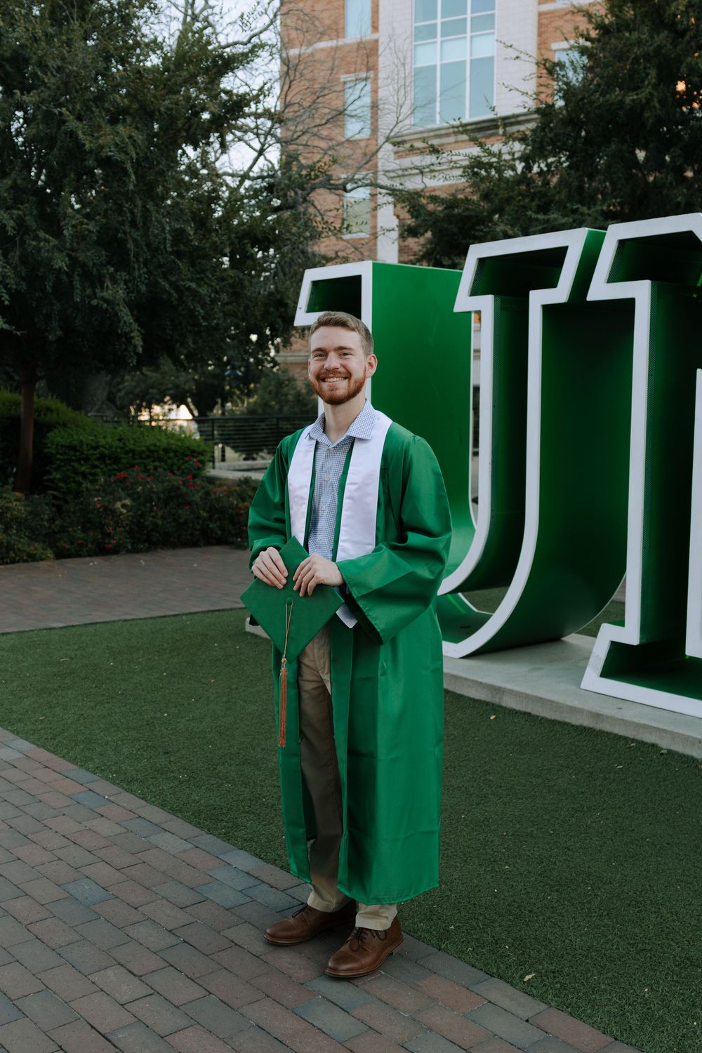 Graduate in green commencement regalia stands proudly by large university sign.