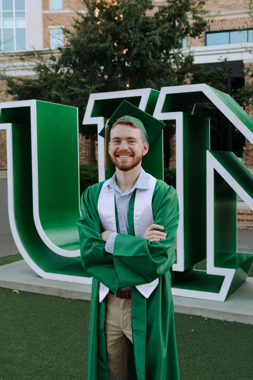 Graduate poses in front of large green university letters wearing traditional commencement regalia.