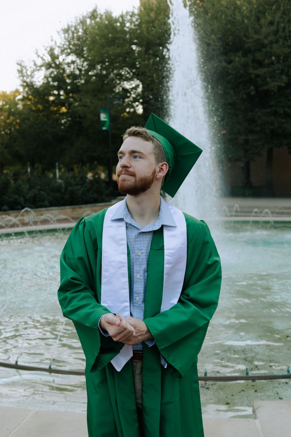 Graduate in green and white regalia poses near a campus fountain during graduation photoshoot.
