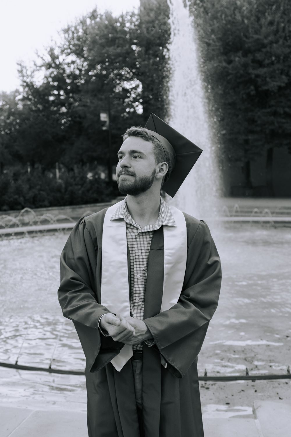 A graduate in cap and gown poses in front of a fountain in black and white photography.