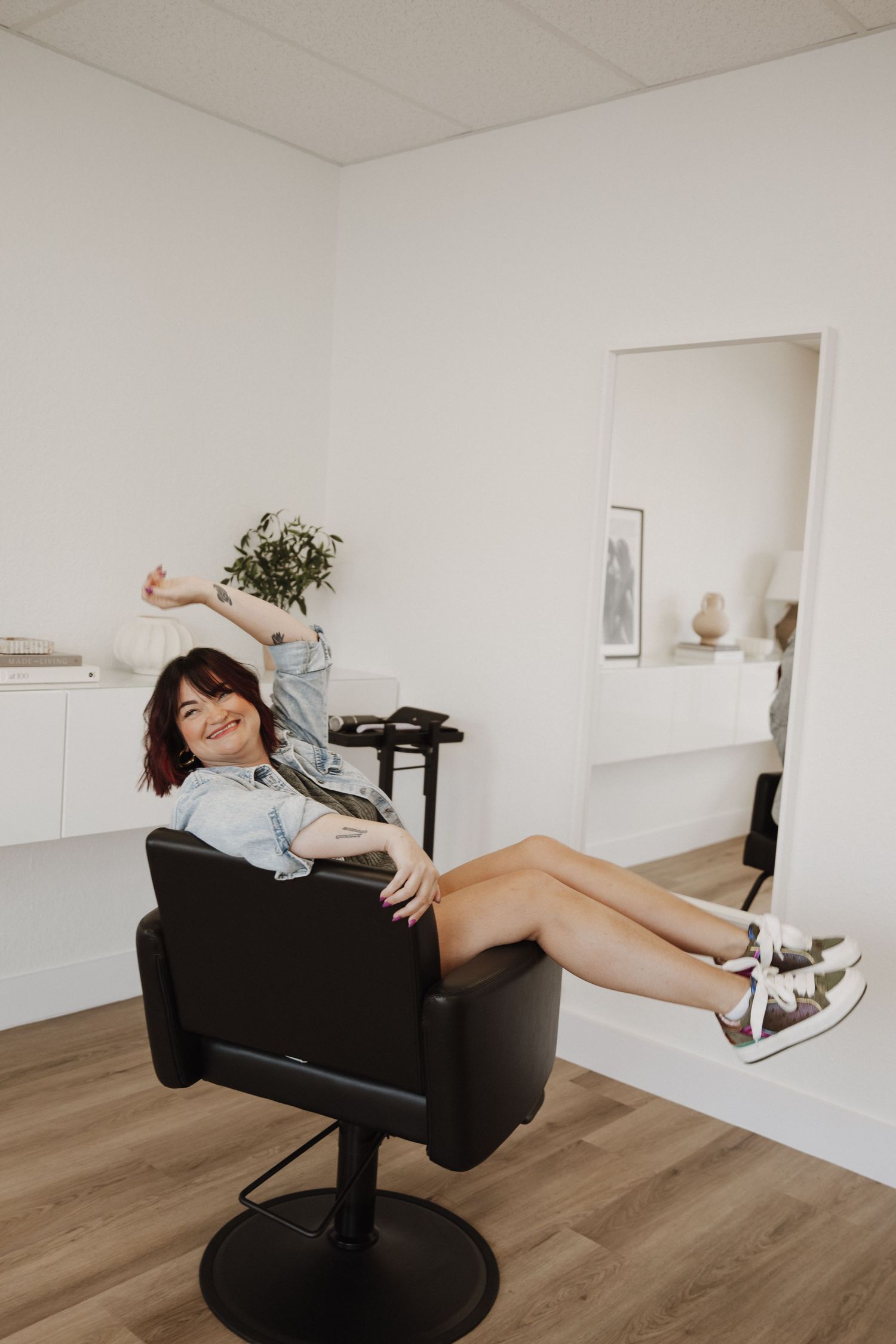 Person lounging and spinning playfully in black salon chair in minimalist white room with wood floors.