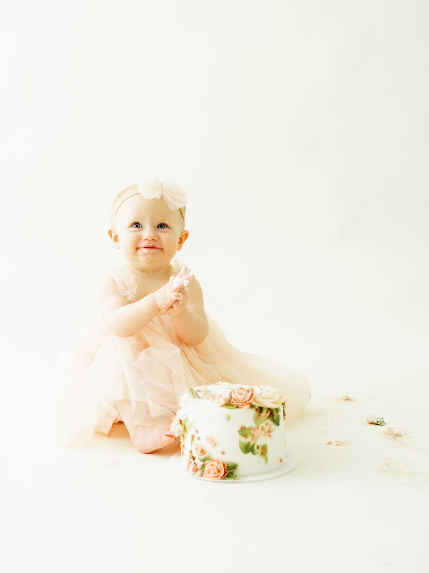 Baby in pink dress sits on white background with small floral cake during cake smash photo session.