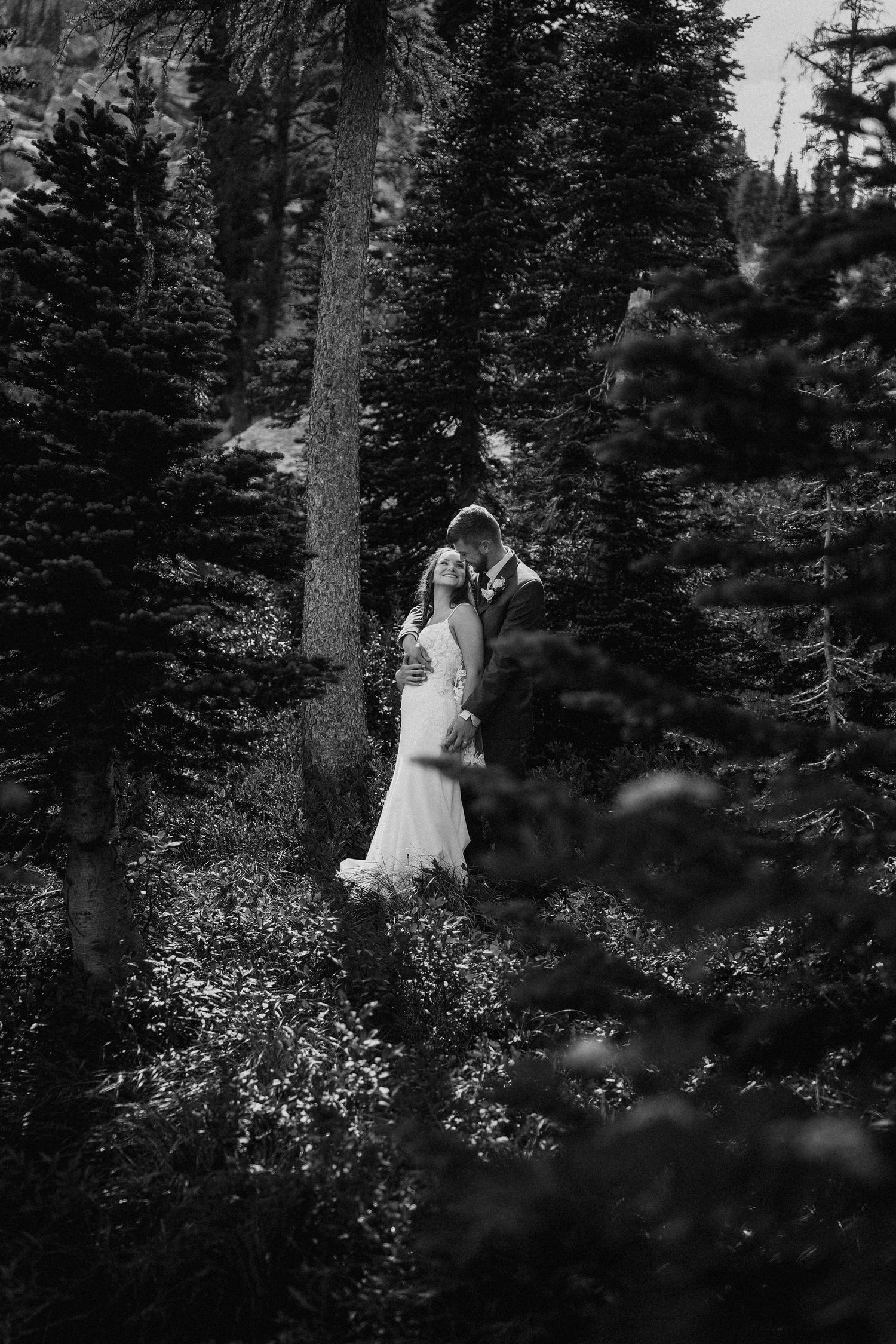 A couple in wedding attire walks through a forest path surrounded by evergreen trees and wildflowers.
