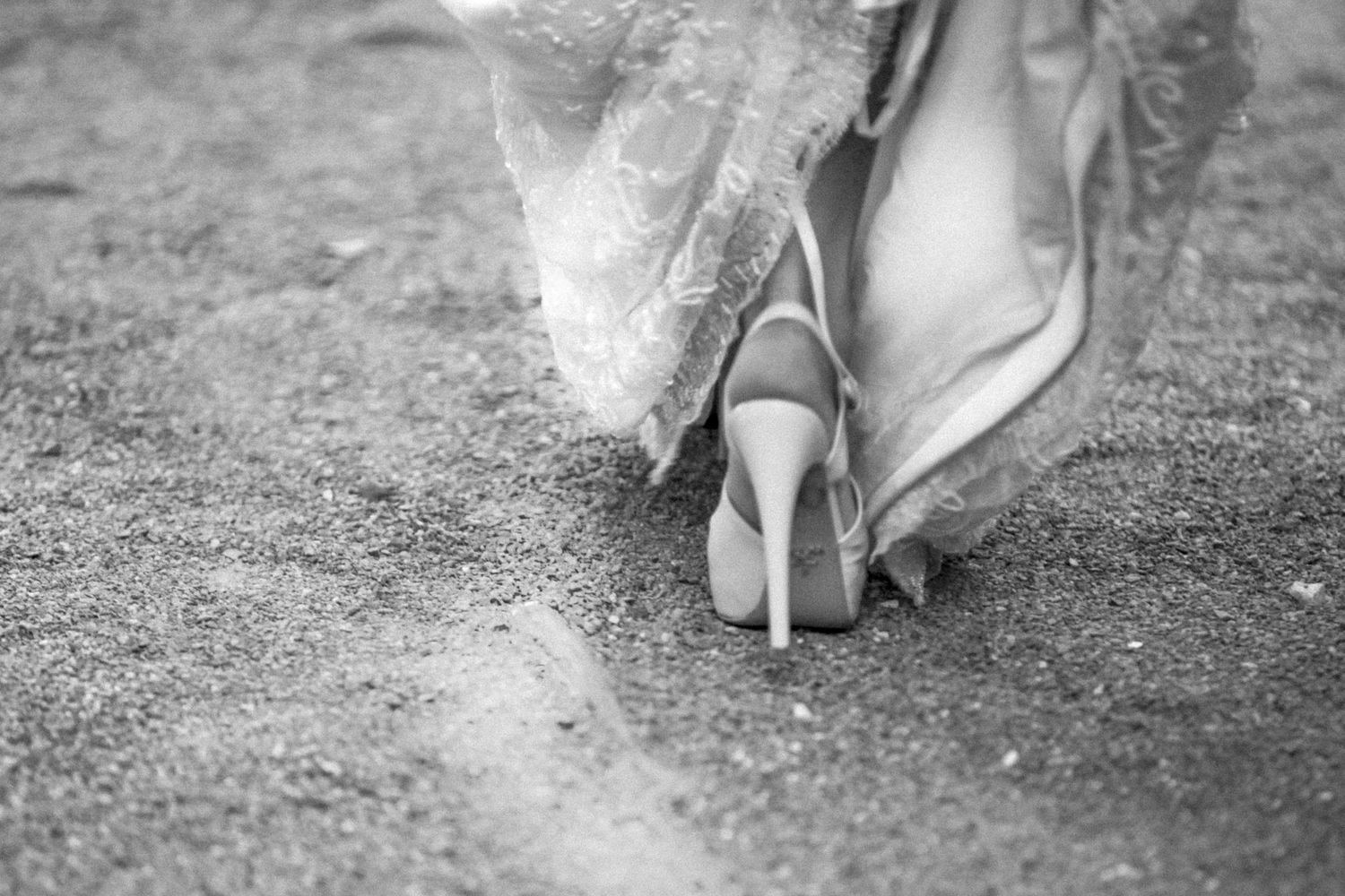 Close-up black and white photo of elegant high heels worn with lace dress against textured ground background.