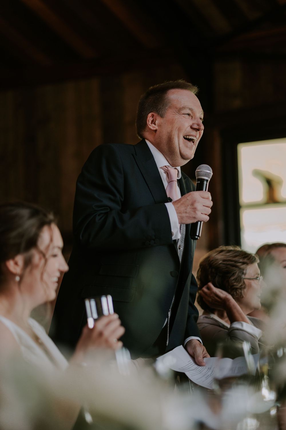 Person in dark suit delivering speech with microphone at wedding reception while guests laugh – Willow Marsh Farm, Loughborough wedding.