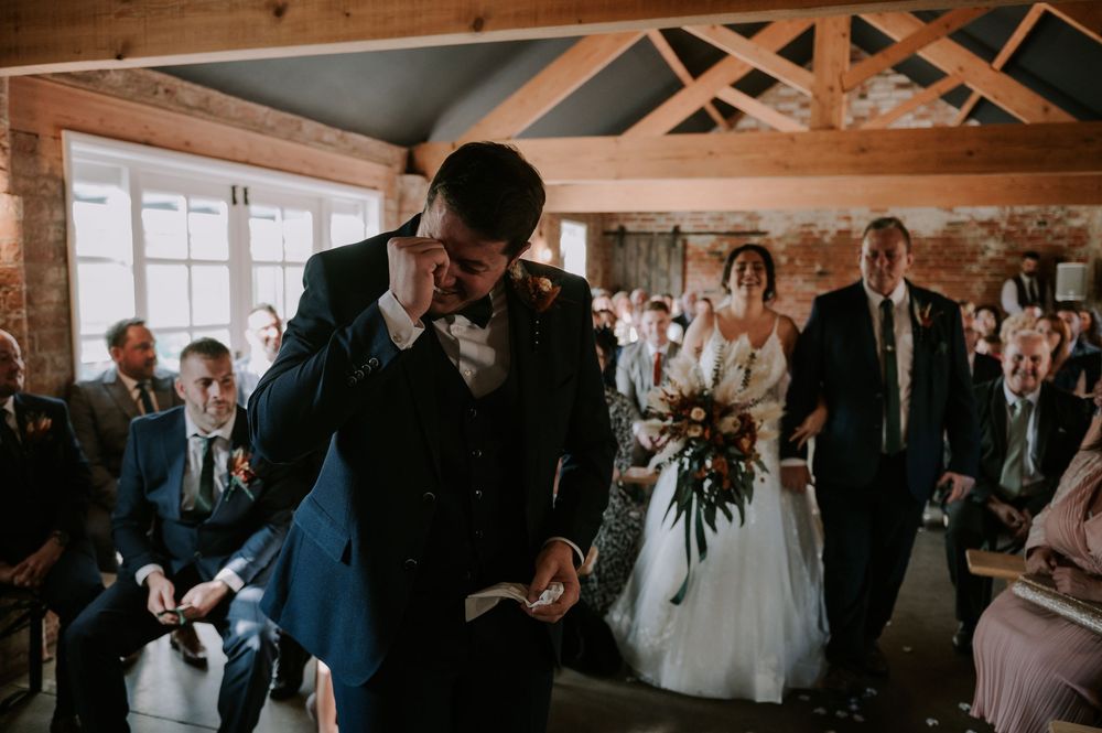 Emotional groom watching as bride walks down the aisle with her father at a rustic barn wedding – Melton Mowbray.