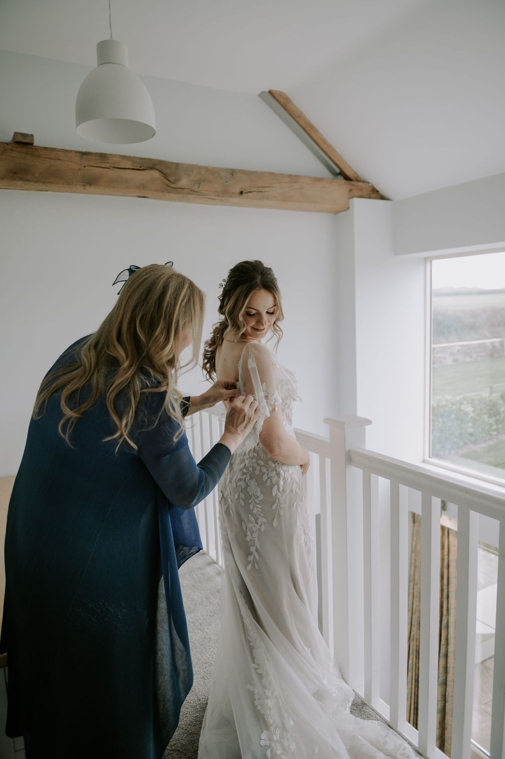 Bride getting her lace gown zipped by her mother in a bright room with wooden beams – Derby wedding