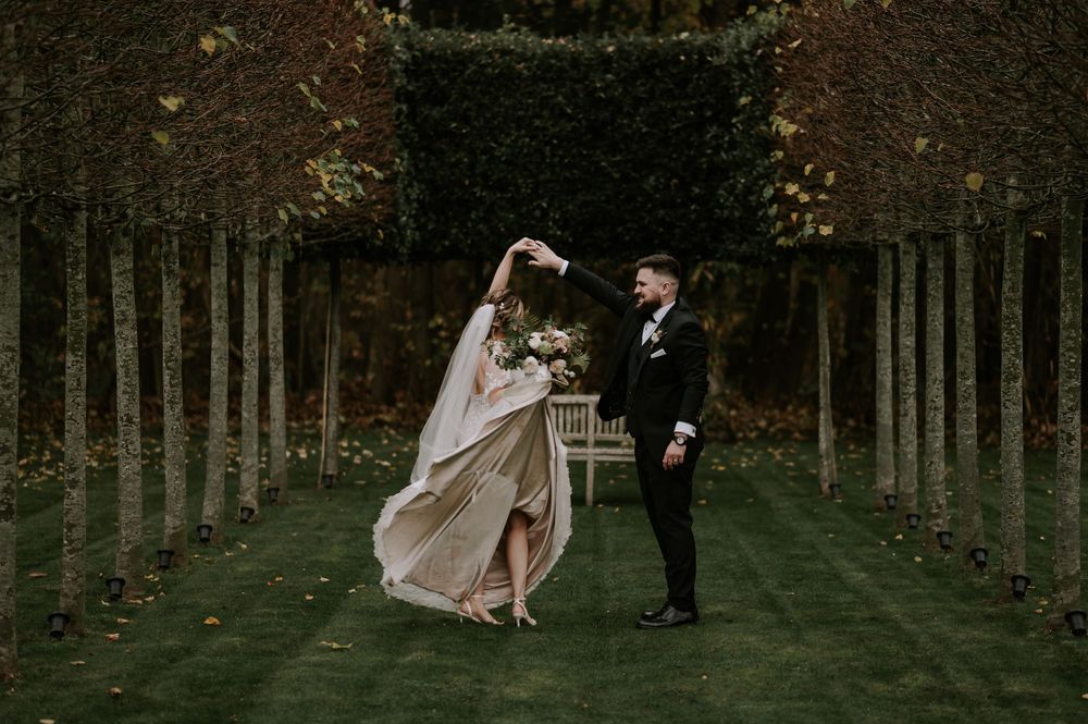 Groom twirling bride in wedding attire on a tree-lined path with autumn foliage – Derby wedding.