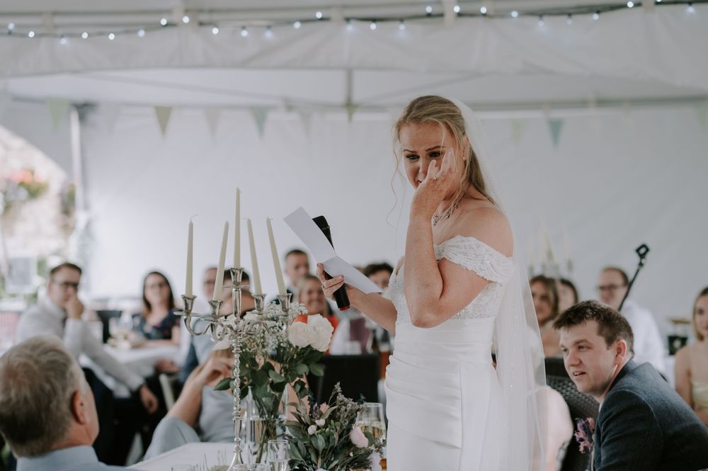 Bride wearing a white wedding dress gives an emotional speech at a reception with string lights and floral centerpieces.
