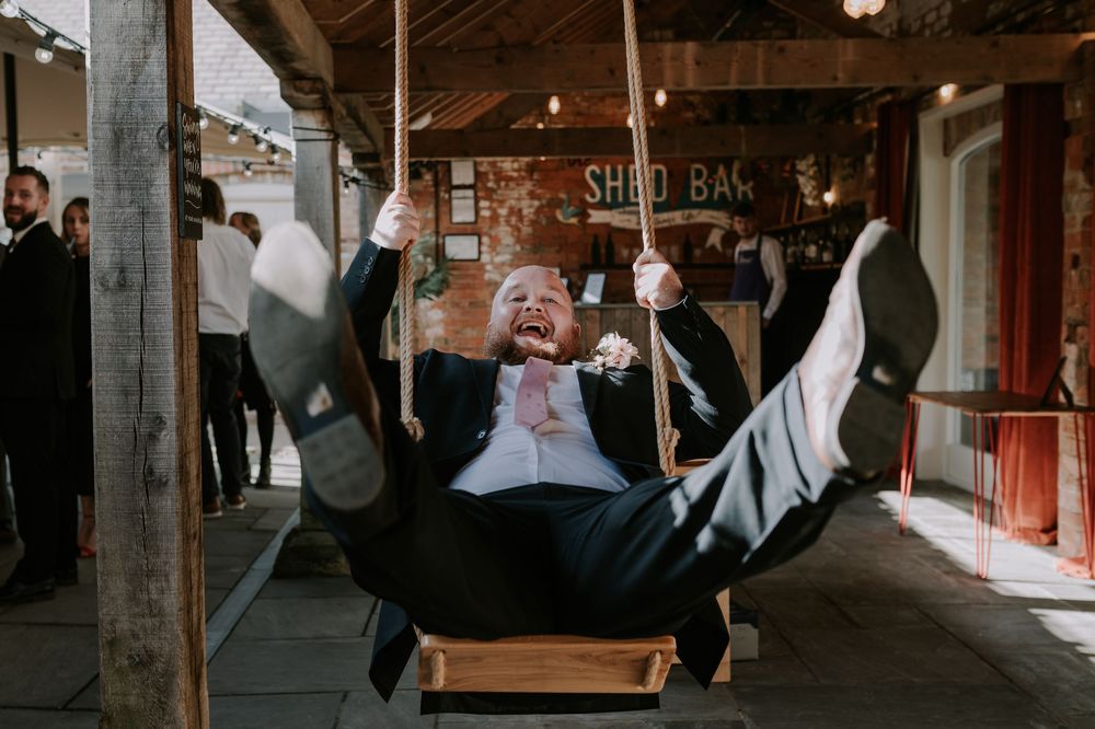 Guest on swing smiling enthusiastically in rustic venue with wooden beams and mural in background – Old Cow Shed, Melton Mowbray wedding