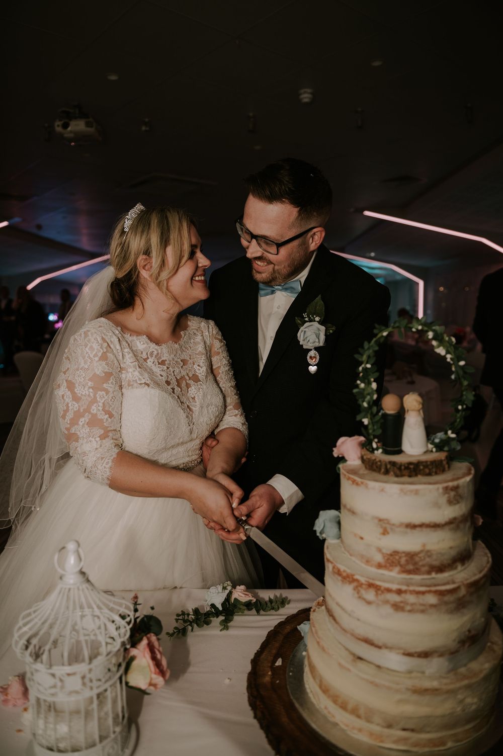 Couple sharing joyful moment cutting rustic semi-naked wedding cake with greenery, reception lights behind – Greetham Valley Oakham wedding.