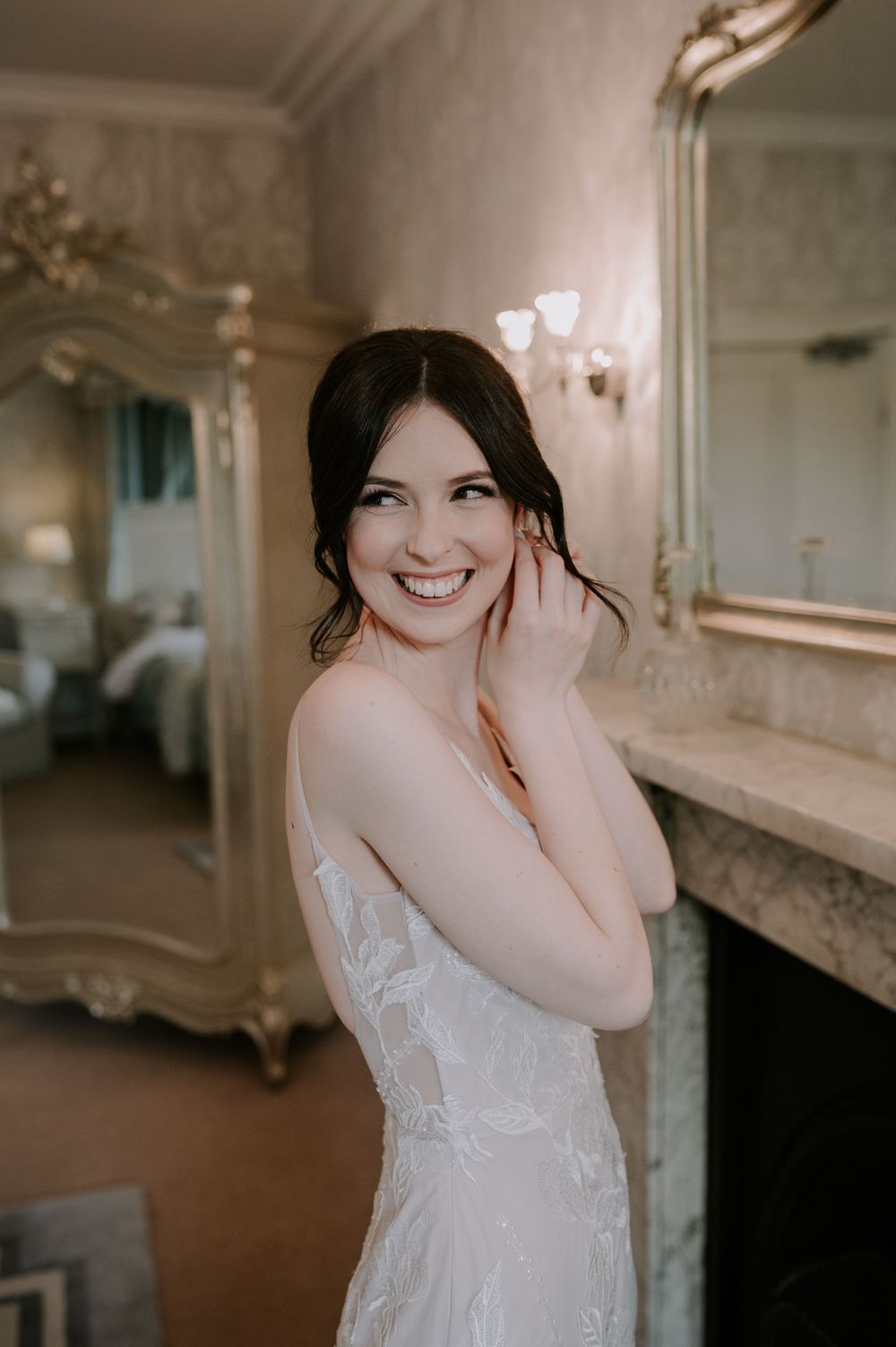 Bride smiling to the left while putting in earrings in elegant bridal prep with lace dress, mirror, and fireplace – Prestwold Hall wedding.