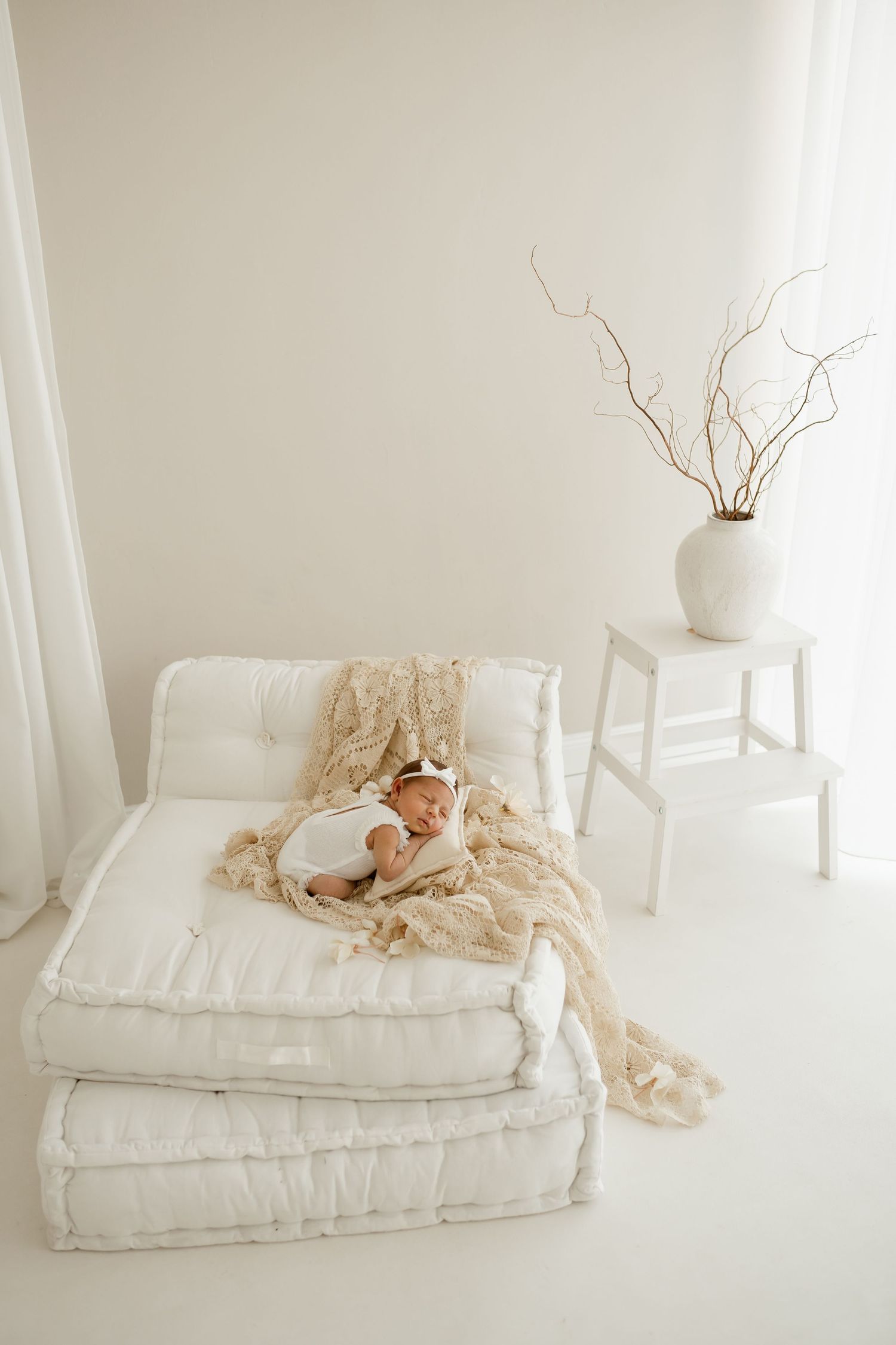 Woman in off-shoulder cream sweater and white pants poses by window in soft natural lighting.