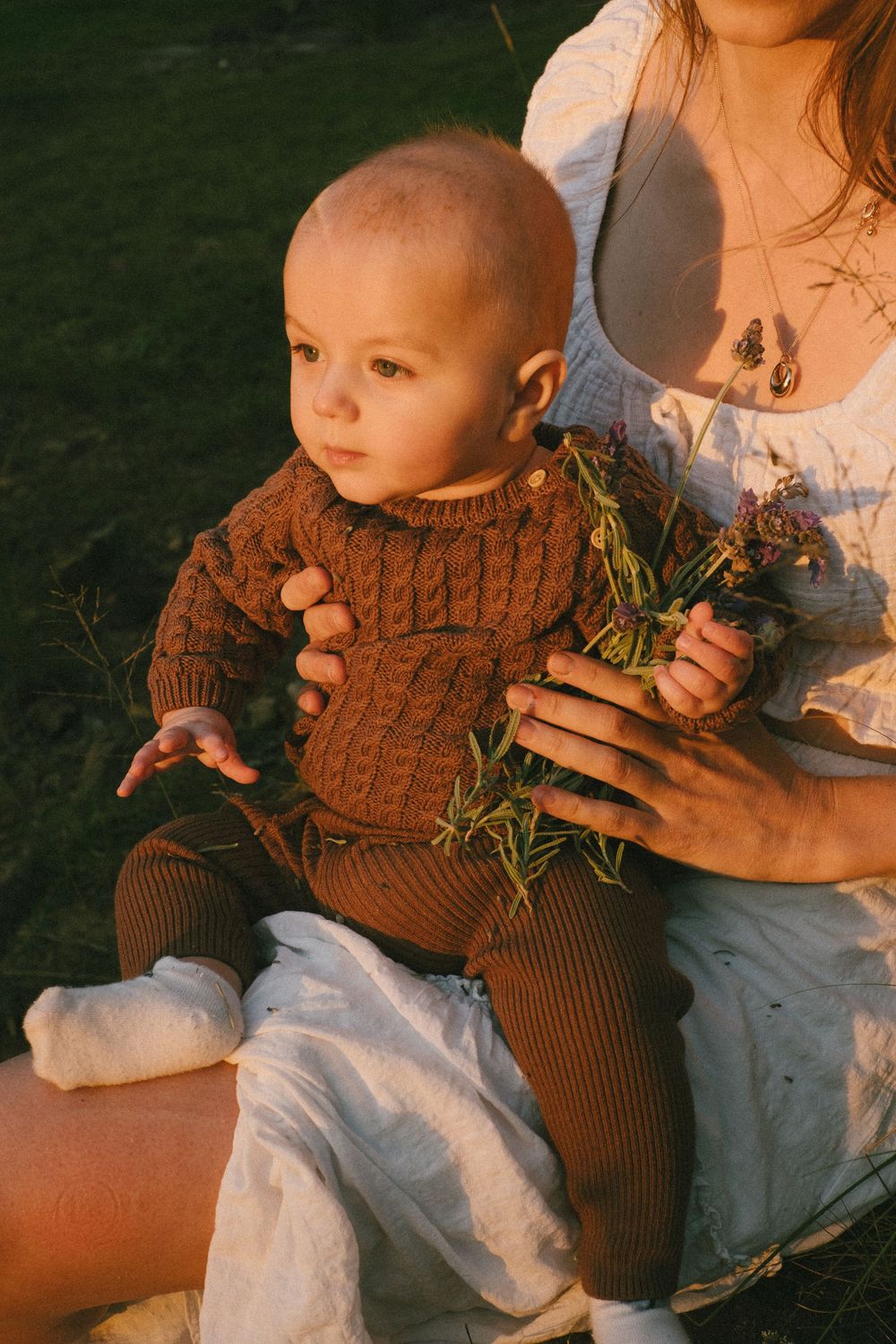 Baby wearing brown knit outfit sits with parent holding wildflowers in warm sunset light.