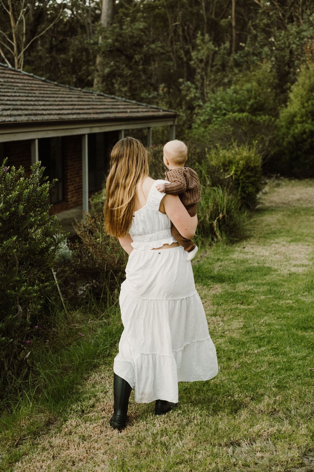 Woman in flowing white dress and boots carries small child in garden setting with rustic house in background.