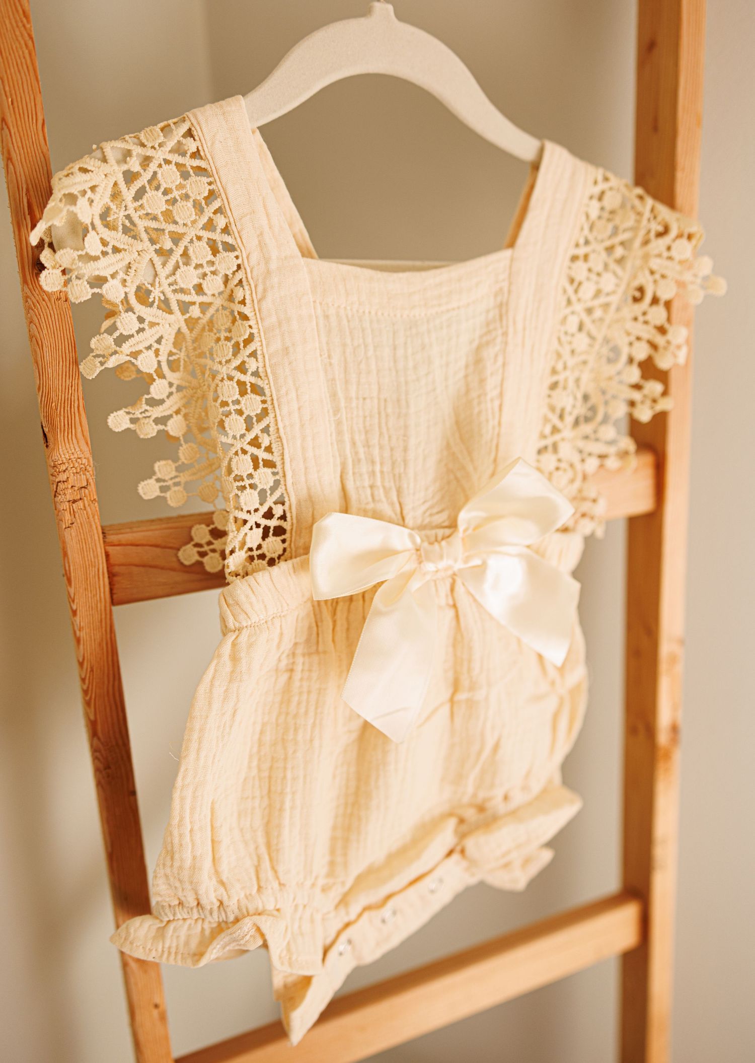 Delicate cream-colored baby romper with lace flutter sleeves and satin bow hanging on wooden ladder.