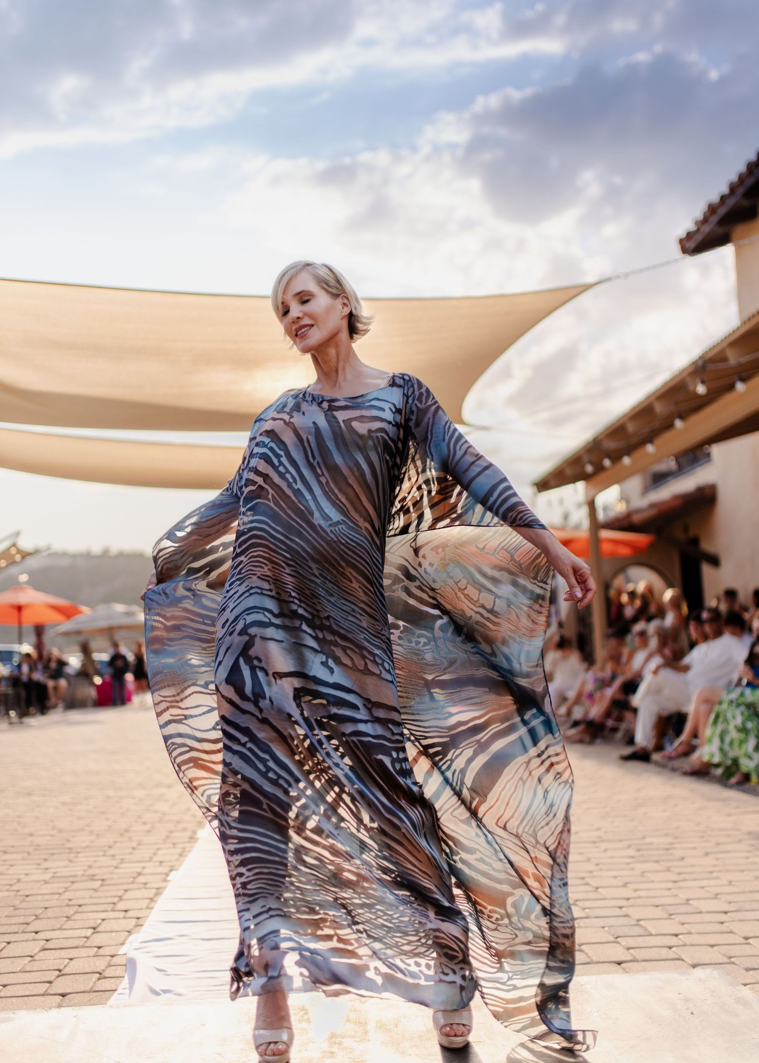 Model in flowing patterned caftan poses under shade sail at outdoor fashion event.