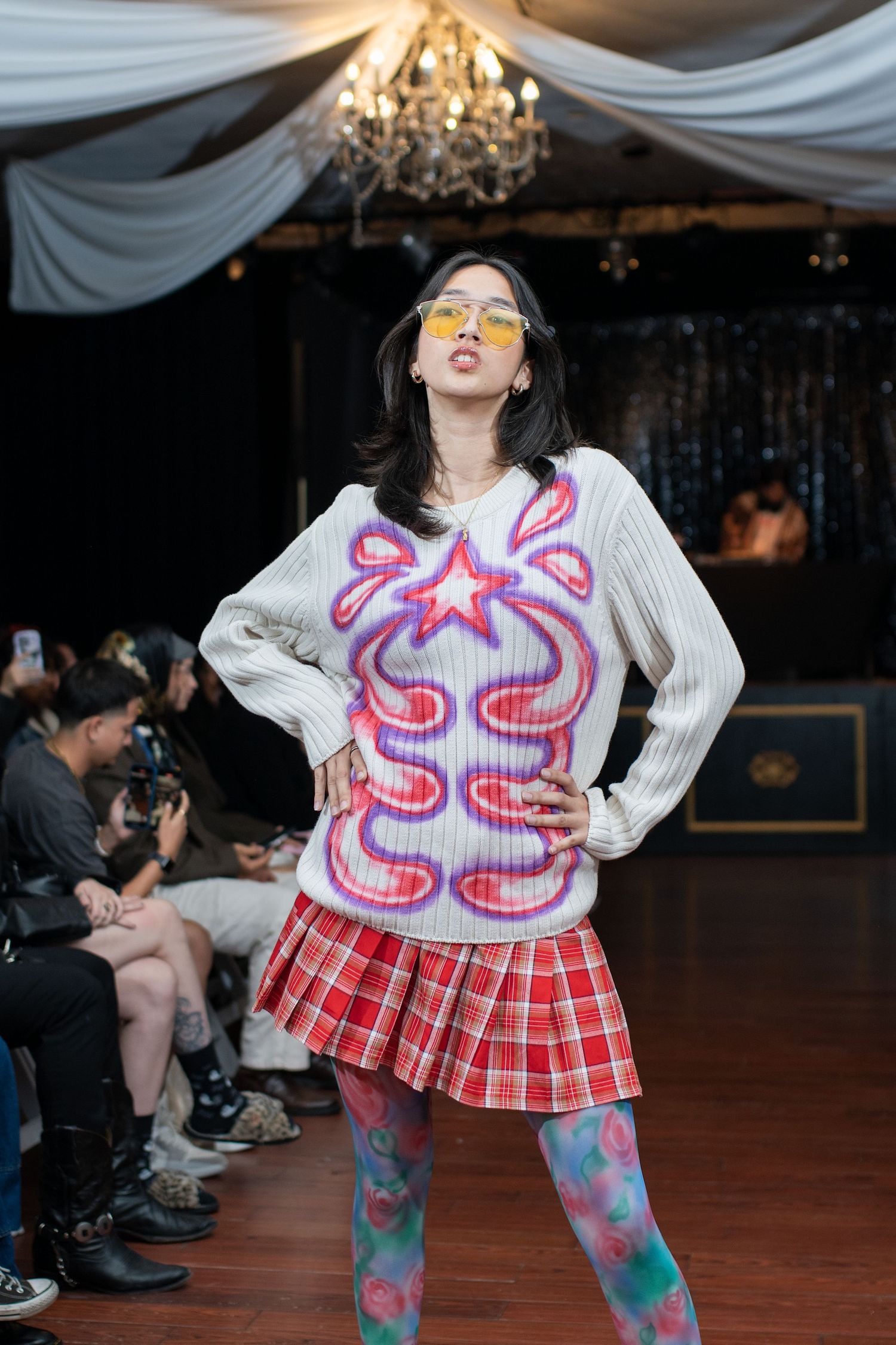 Model struts in playful pink butterfly sweater with plaid skirt and patterned tights on indoor catwalk.