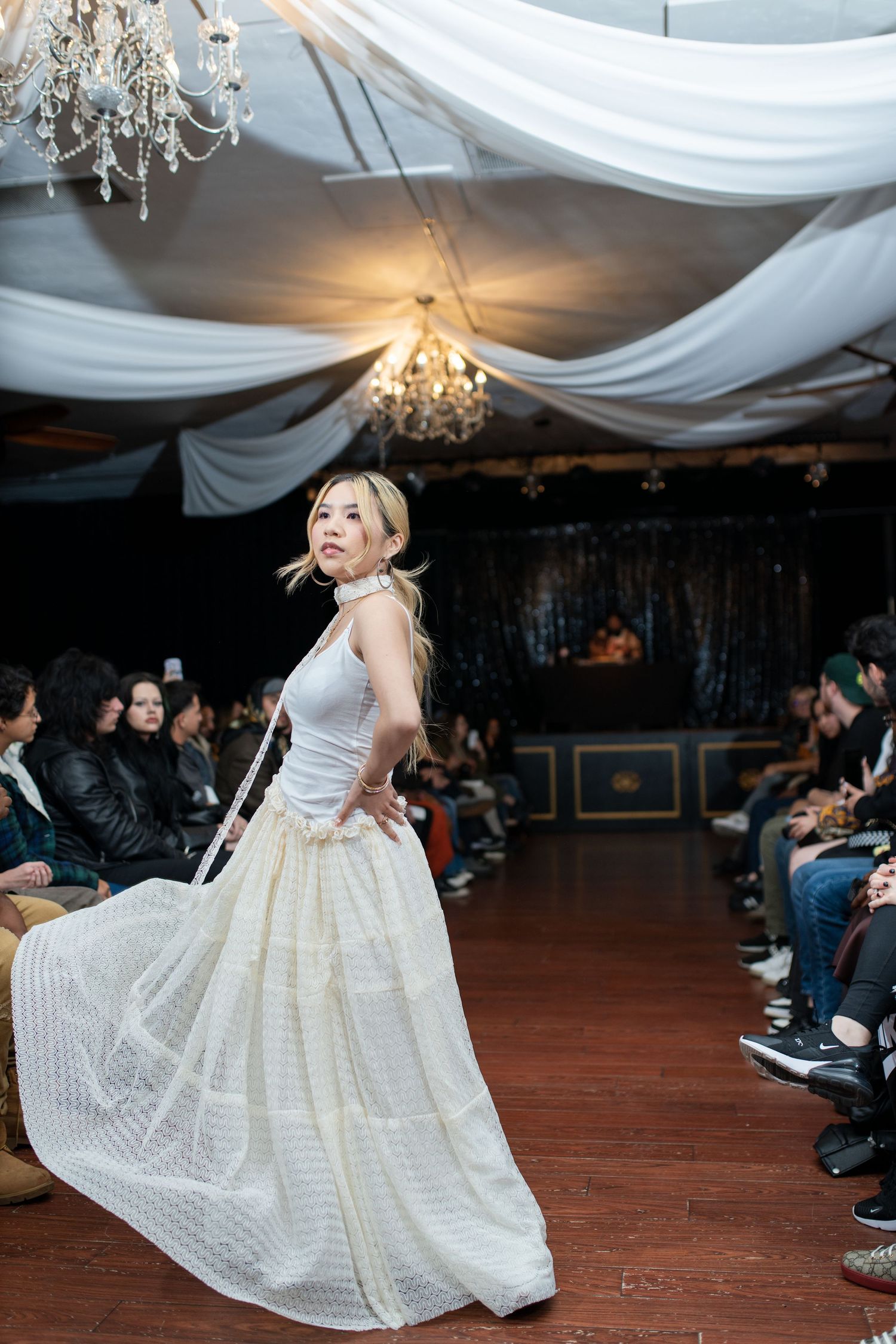 Model twirls in white vintage-inspired gown on runway under draped fabric and chandeliers.