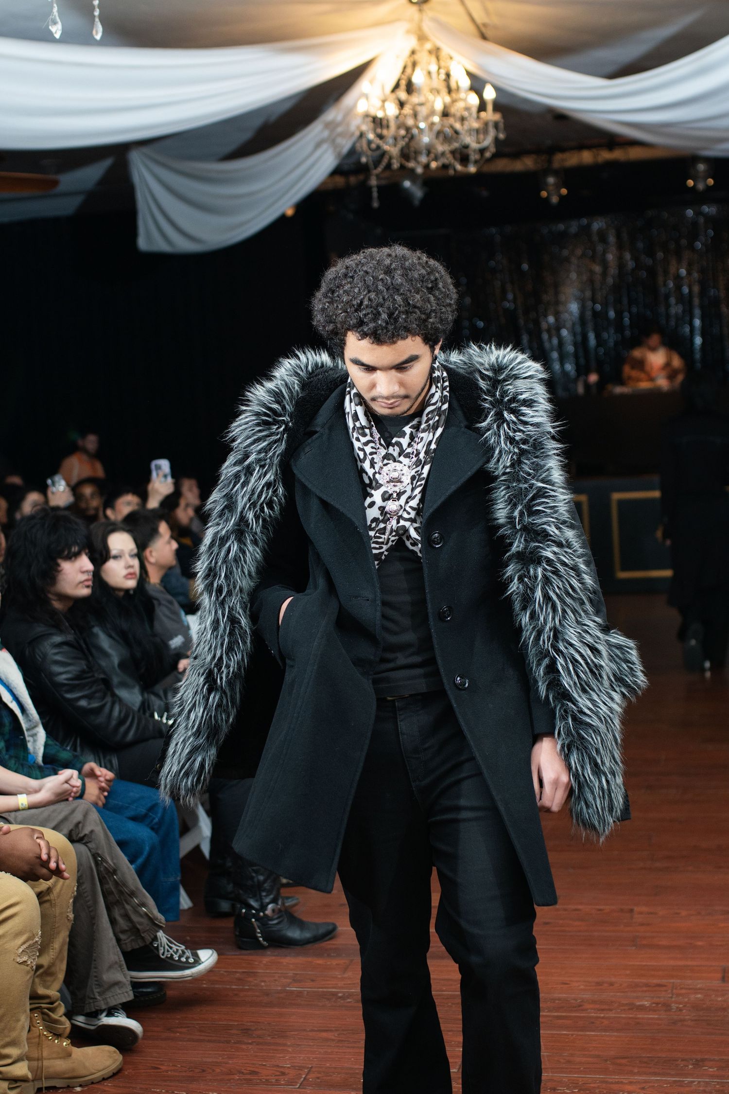 Model presents black coat with dramatic fur trim during indoor fashion show.