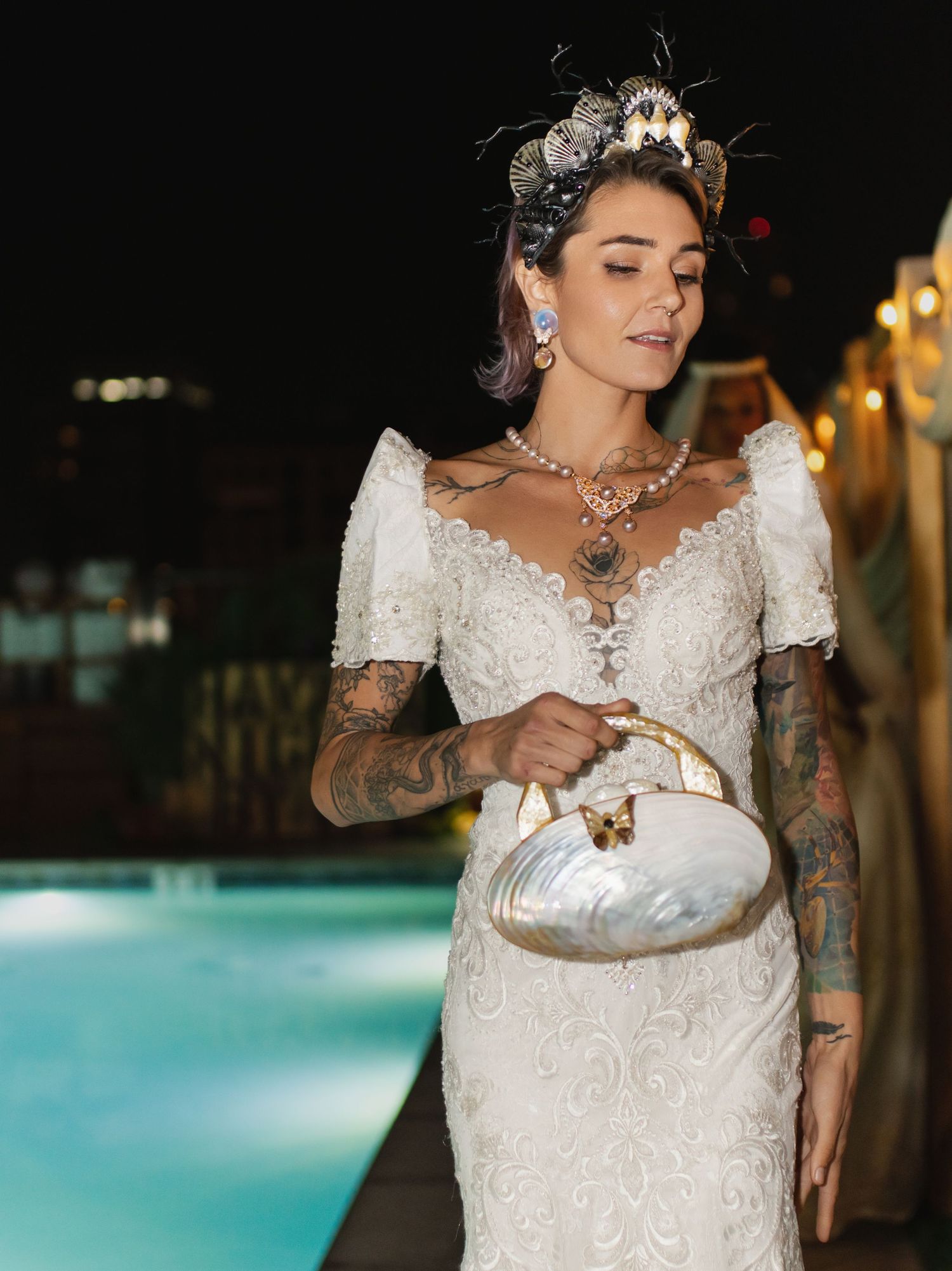 Elegant model in white lace dress with tattoos poses by pool holding vintage purse.