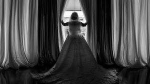 A silhouetted figure in a flowing ballgown stands at a window framed by dramatic floor-length curtains in black and white.