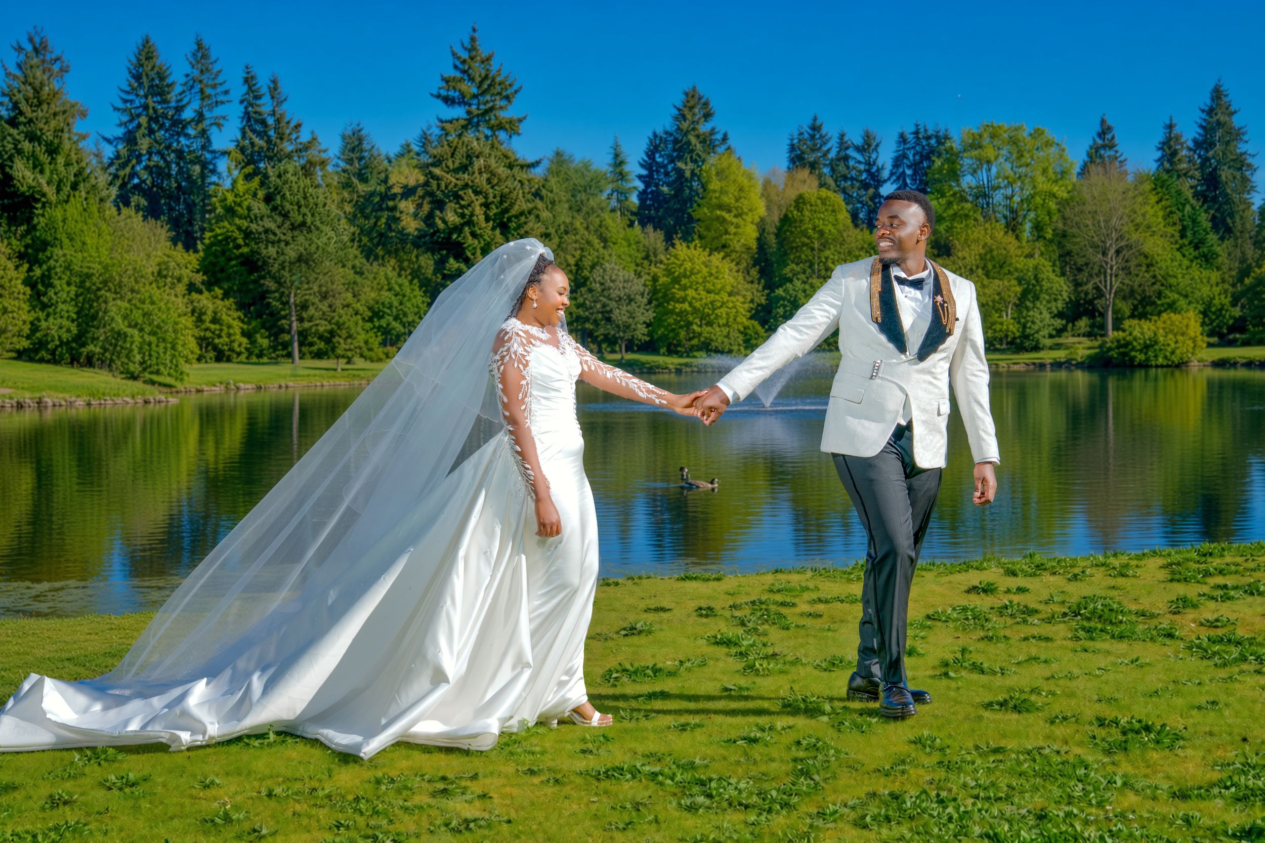Bride and groom holding hands by a serene lake surrounded by lush green trees on a sunny wedding day.
