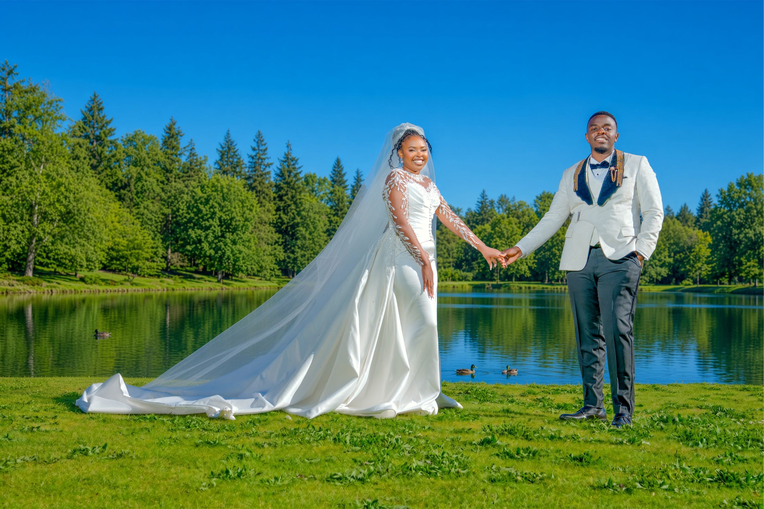 Bride and groom hold hands by a serene lake surrounded by lush green trees on a sunny wedding day.