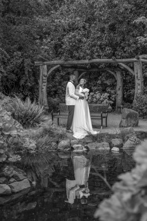 Black and white wedding photo of couple embracing by pond with wooden arbor and garden reflection.