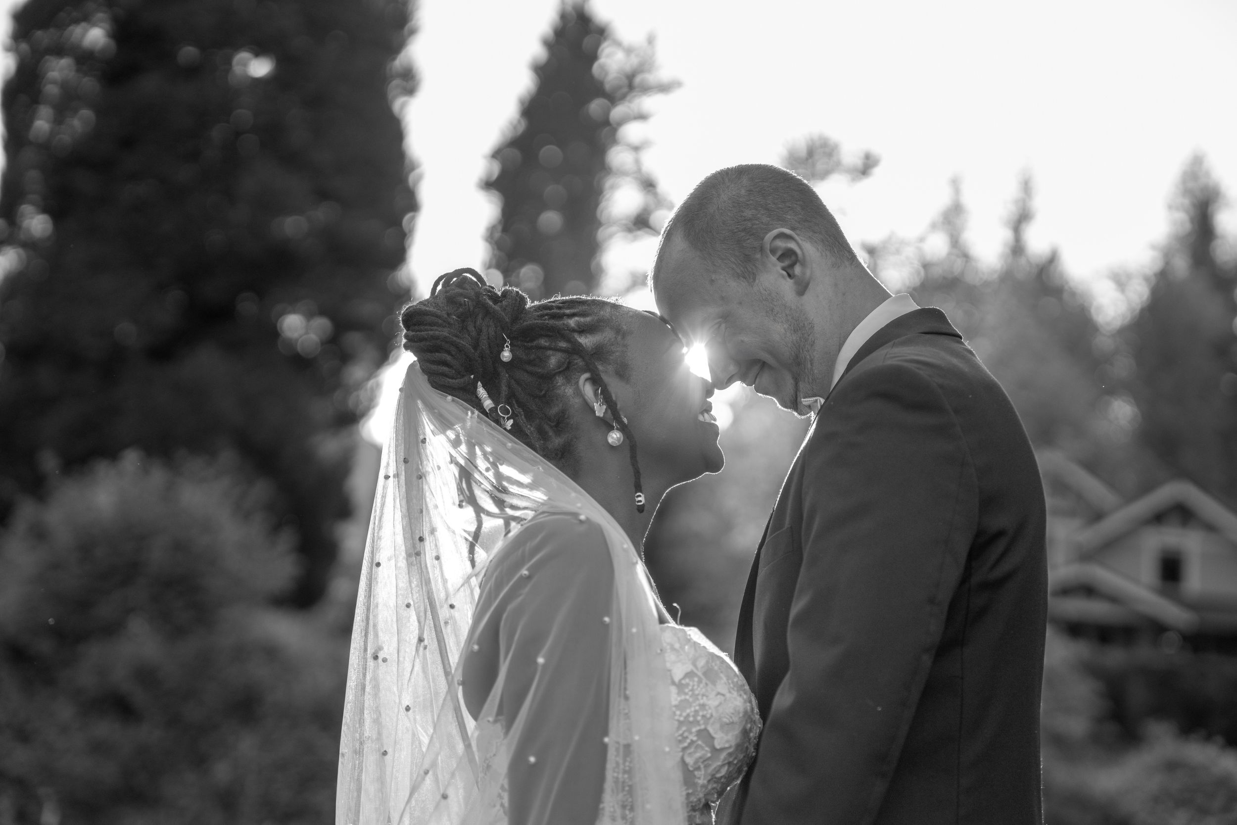 Black and white wedding photo of bride and groom touching foreheads outdoors, veil flowing, trees in background.