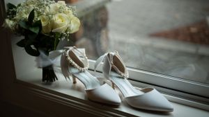 Elegant white wedding shoes and bridal bouquet of cream roses displayed on windowsill in natural light.
