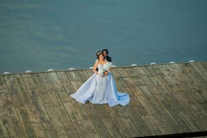 A couple in wedding attire dances on a wooden pier overlooking water.