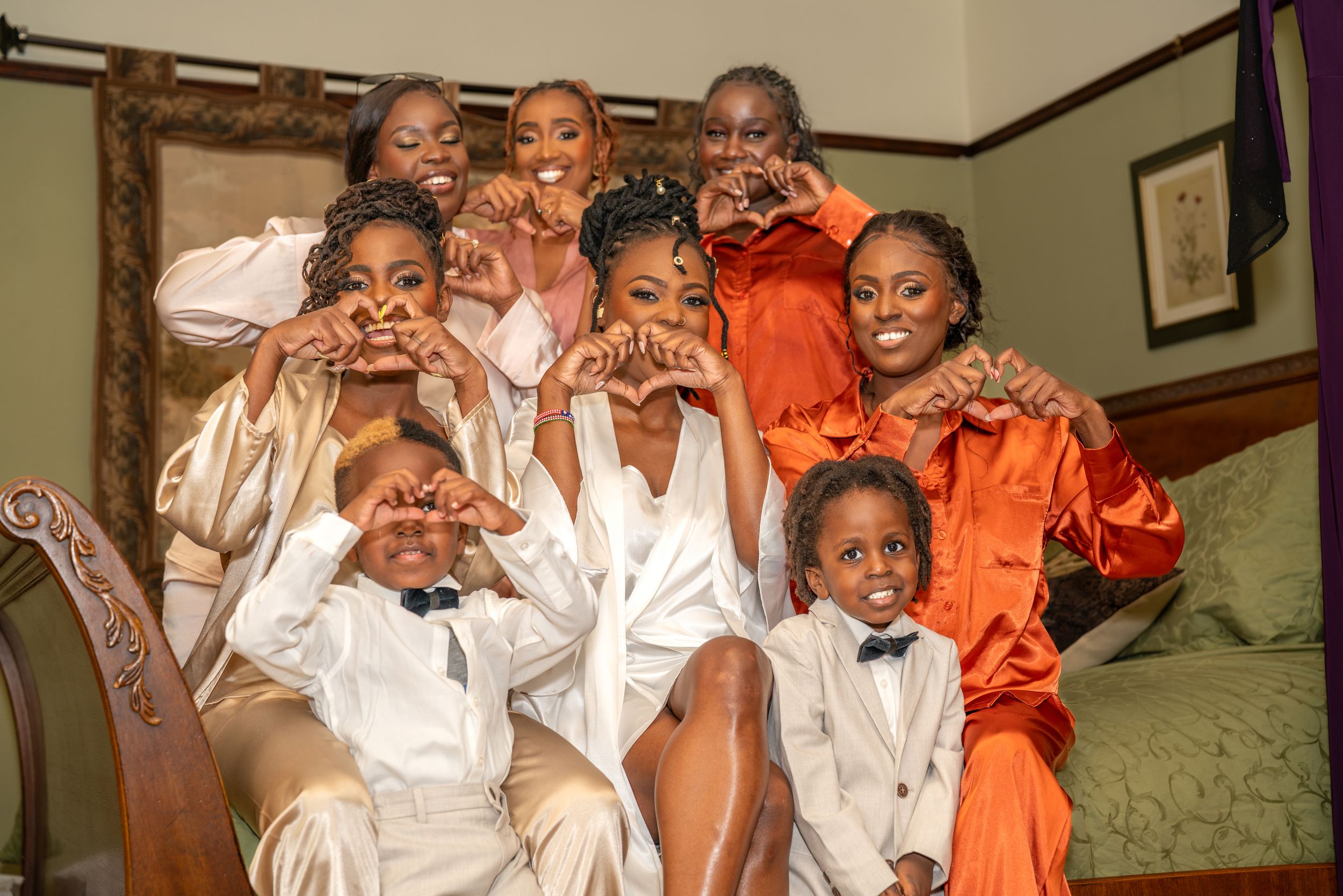 Group of joyful women and children posing together, making heart shapes with hands, dressed in satin and orange outfits.
