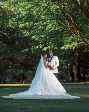Elegant couple in wedding attire poses gracefully on lush green lawn surrounded by trees during sunset photoshoot.