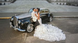 A vintage car photoshoot with a flowing white wedding dress trailing elegantly on the ground.
