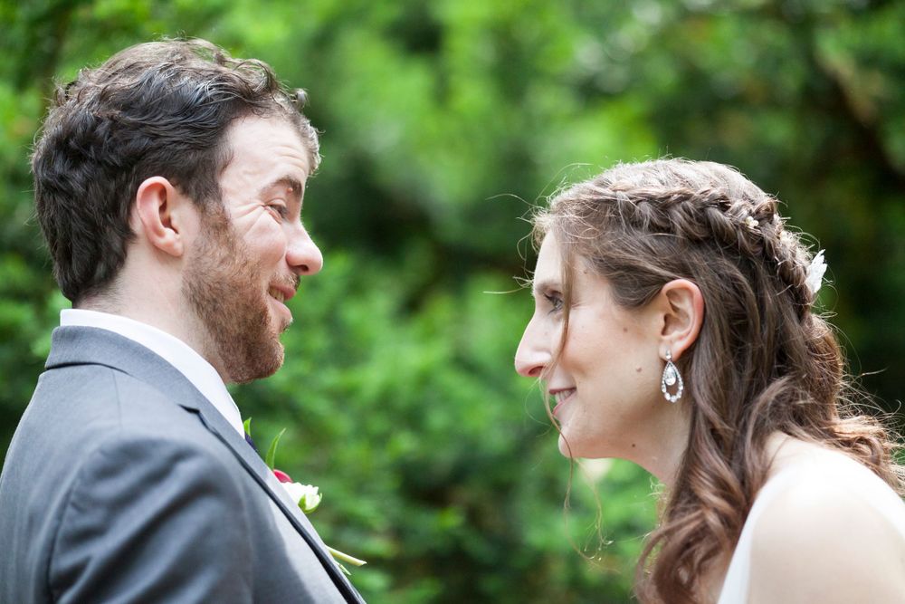 Bride and groom share intimate smile face to face surrounded by lush green foliage.