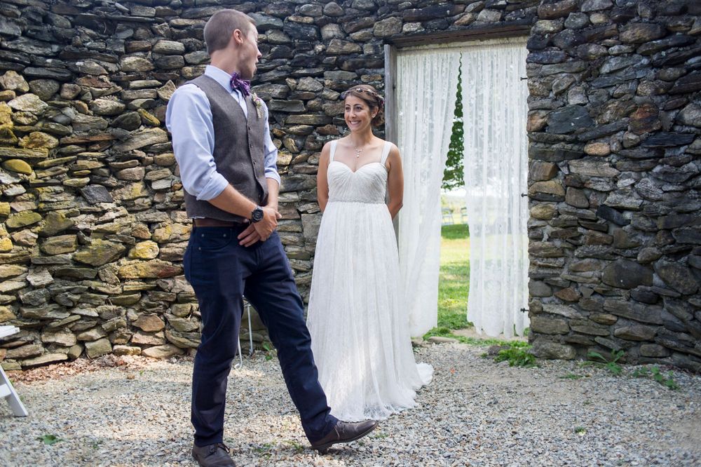 Groom turns to see bride in white gown during first look against rustic stone wall.