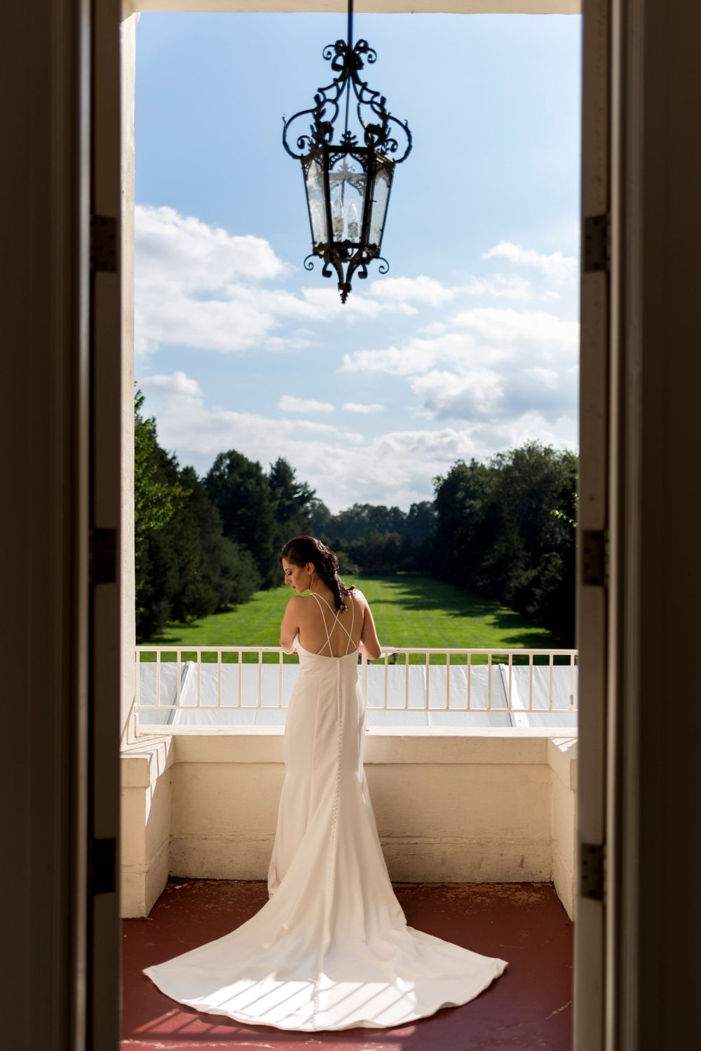 Bride in elegant white gown stands on balcony overlooking manicured green estate grounds.