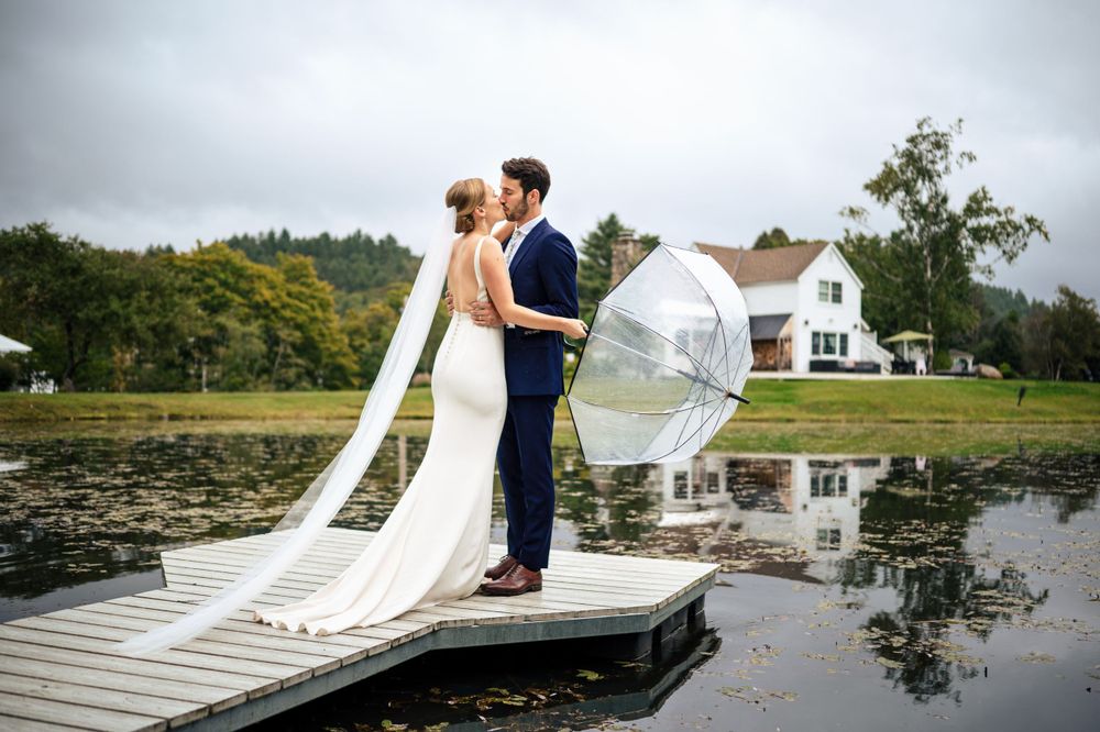 Bride and groom kiss on dock by tranquil lake with clear umbrella on overcast day.